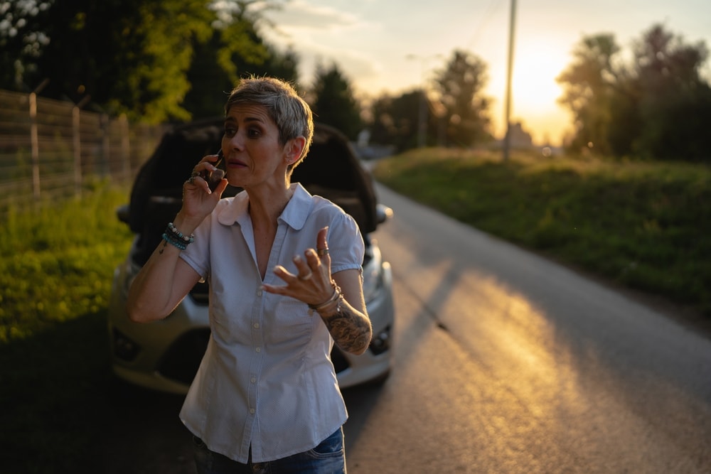 mature female standing on the road in the evening sunset by the broken vehicle making a phone call for roadside assistance towing service